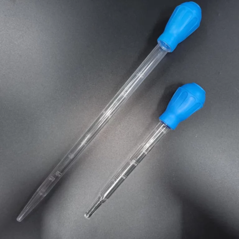 Two clear plastic dropper/pipette tools with blue bulb caps lying on a dark surface, used for measuring and transferring liquids.