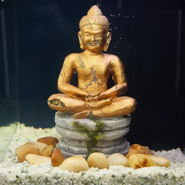 Small golden Buddha statue meditating on stacked rock pedestal, surrounded by white pebbles and stones against a dark background